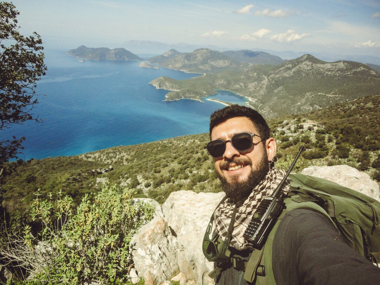 Hadi Yürüyelim profesyonel dağ rehberi Şükrü Aydın, Fethiye Ölüdeniz manzarasına karşı trekking ekipmanları ve telsiziyle gülümsüyor.