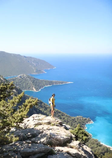 Likya Yolu Yediburunlar parkurunda, yüksek bir kayalık üzerinden turkuaz deniz ve koy manzarasını izleyen Hadi Yürüyelim rehberi.